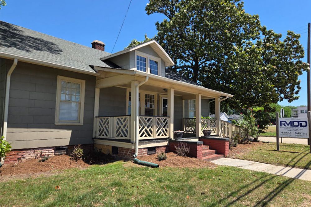 Charming gray house with wooden porch and lush trees under a bright sky, featuring a real estate sign.